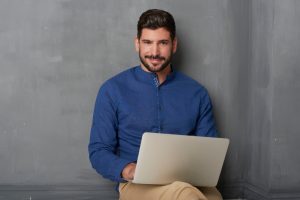 גבר יושב עם הגב אל הקיר ועל רגליו מונח מחשב נייד.